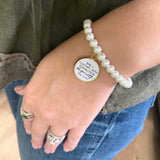 A natural pearl beaded slider bracelet with an 18K gold-plated chain and a dangling glass charm with the words "She is Clothed with Strength & Dignity"
